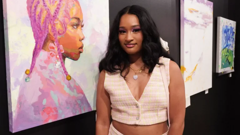 A person stands in front of a colorful portrait painting on a gallery wall, wearing a sleeveless outfit, amid other abstract artworks displayed in the background.