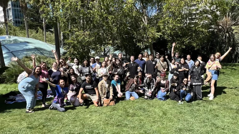 A group of people stands waving on a grassy area, surrounded by trees and skyscrapers, under a clear blue sky.