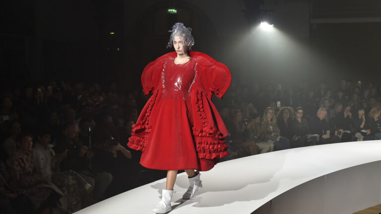 Model wearing a voluminous red dress with ruffled details and white boots walks on a curved runway. Audience seated in the background, and overhead lights illuminate the scene.