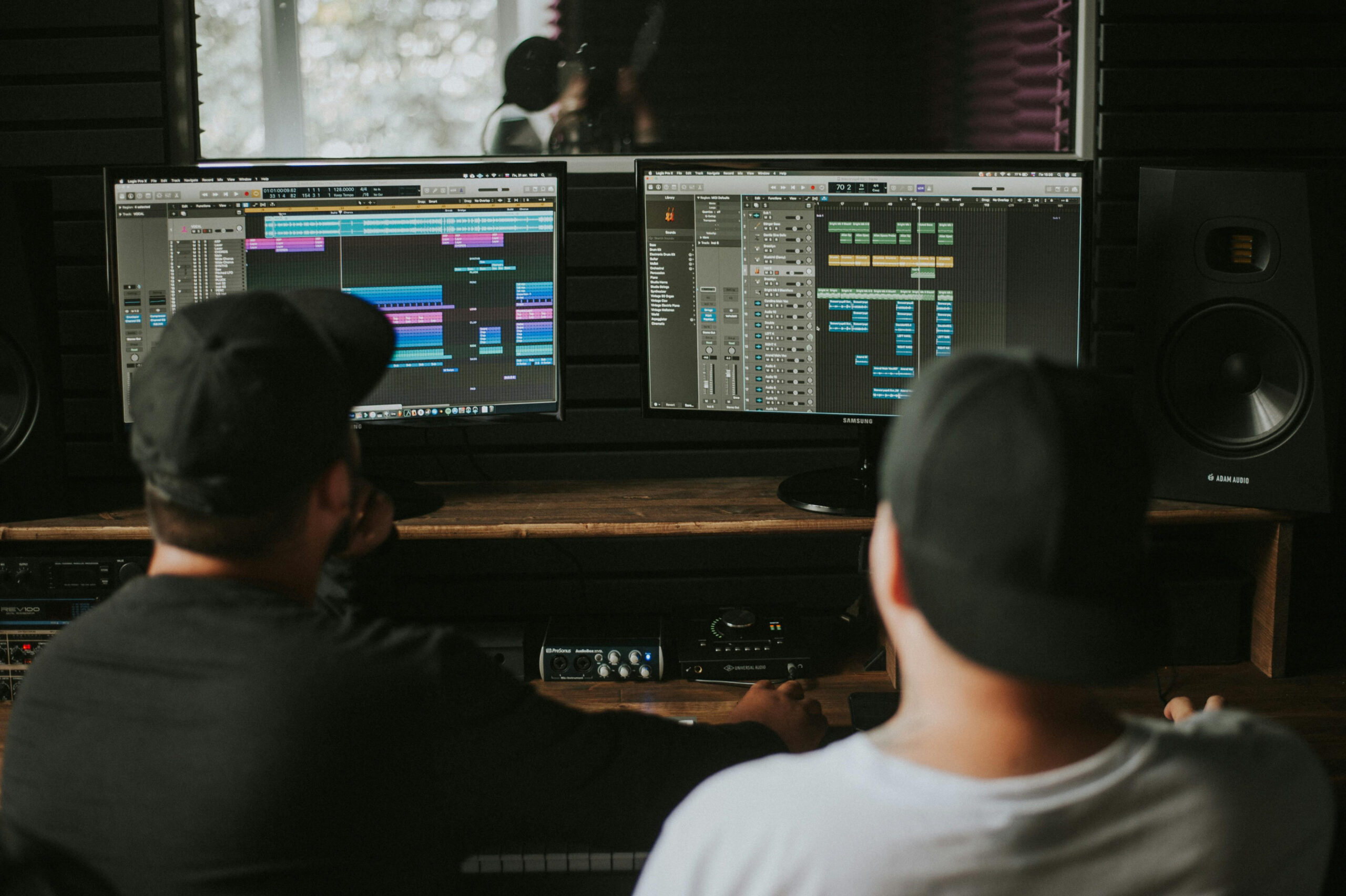 Two people in black caps work on music production at a studio, facing dual monitors displaying digital audio workstations, surrounded by speakers.