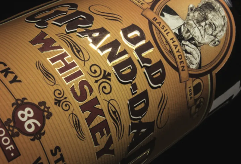 A whiskey bottle label displays "Old Grand-Dad Whiskey" with "86 Proof" beneath. The background features ornate designs and a portrait labeled "Basil Hayden." The label is angled against a dark background.