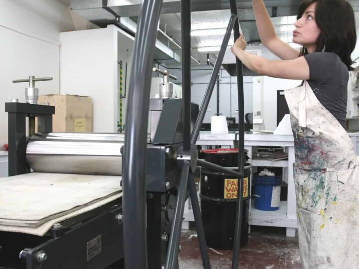 A person wearing a paint-stained apron operates a large printing press wheel in an art studio. Tables, tools, and equipment surround them, with visible pipes on the ceiling.