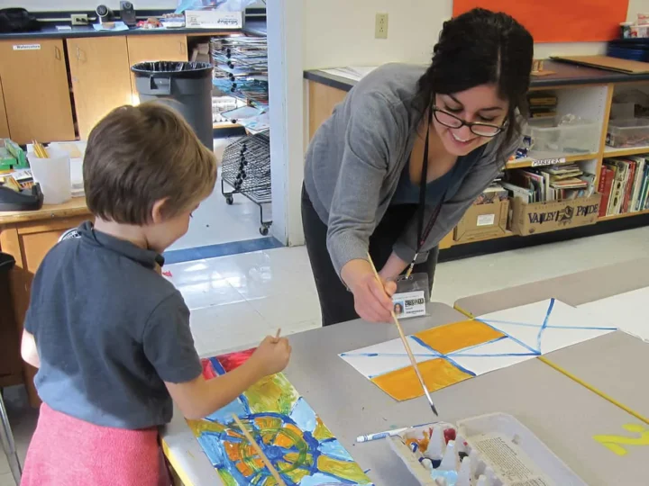 A teacher and child paint on large paper at a classroom table. Art supplies and books are in the background. The teacher wears an ID badge, and both are smiling while working together.