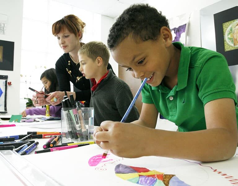 Children draw at a table with art supplies while a teacher observes. The classroom features art on the walls, suggesting a creative, educational environment.