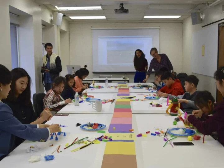 Children and adults are engaged in arts and crafts at a long table, using colorful materials. The room has a projector screen and bright lighting, creating a collaborative classroom environment.