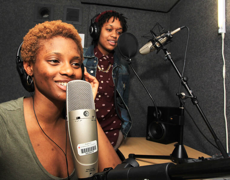 Two people wearing headphones speak into microphones in a recording studio. The room has soundproof walls, a desk with audio equipment, and paperwork on the wall.