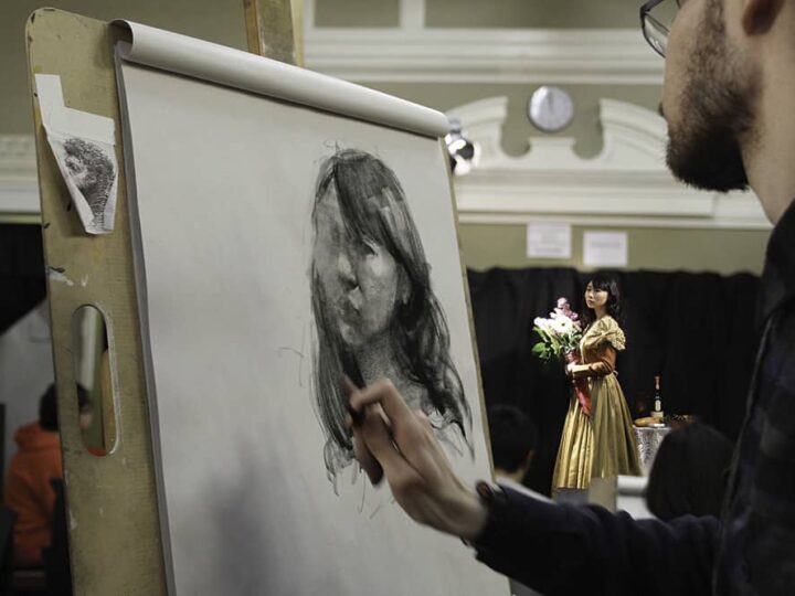 An artist sketches a portrait on paper while observing a model posing in a gold dress with flowers, set in a classic studio environment.