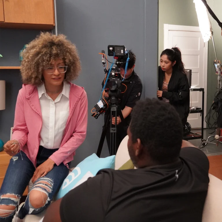 A behind-the-scenes view of a film set where a woman in a pink jacket and glasses talks to a man sitting on a couch. Behind them, a camera operator films the scene while another crew member watches from a doorway. The setting appears to be a cozy living room with lighting and film equipment visible in the background.