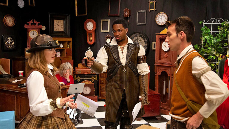 Three people in period costumes discussing in a room filled with clocks and picture frames.