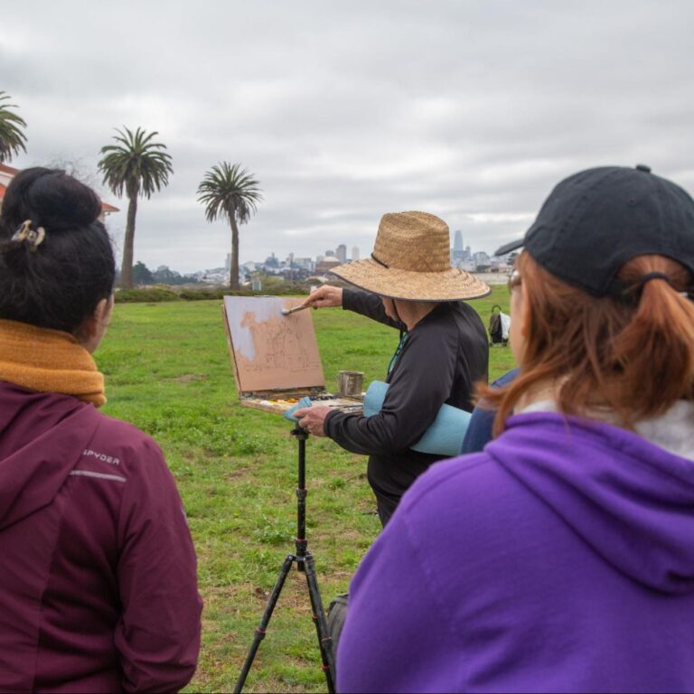 Several people watching as a person in a straw hat is painting on an easel outdoors