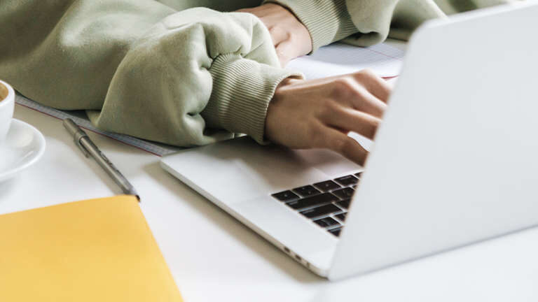 A person working on a laptop at a white desk, with a cup of coffee, a smartphone, a pen, and some notebooks and papers around.