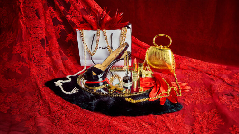 Luxury accessories and cosmetics displayed on red lace fabric, including a Chanel shopping bag and high heels.