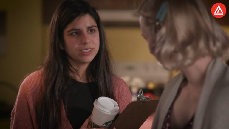 A woman with dark hair holding a coffee cup talks to another woman with gray hair, who is writing on a clipboard.
