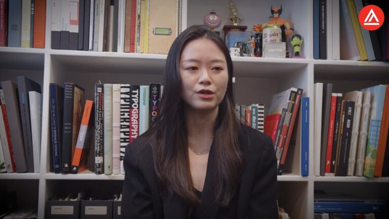 Woman speaking in front of bookshelf filled with books and collectibles.