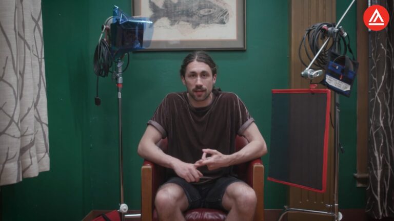 Man sitting in a chair surrounded by filming equipment in a room with green walls.