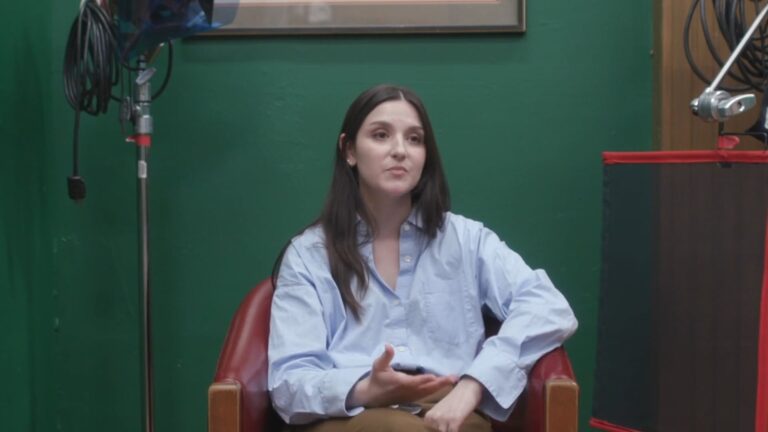 A woman with dark hair sitting in a red chair, holding a phone, in front of a green wall.