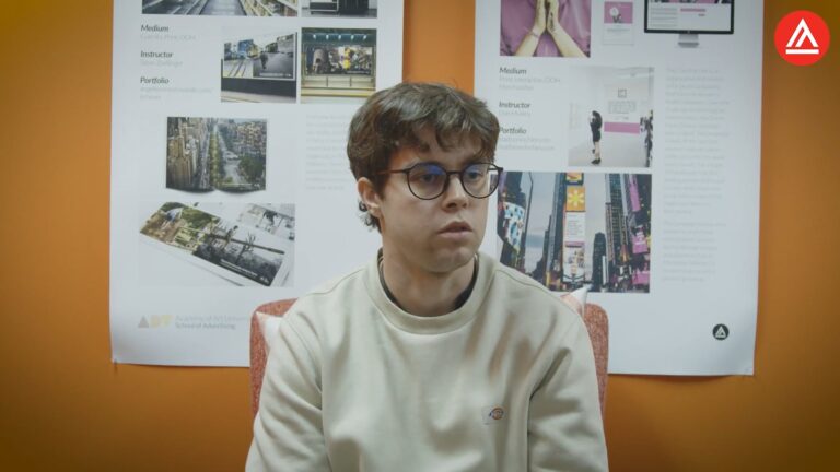Young man with glasses sits against an orange wall with posters behind him.
