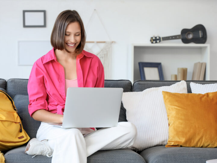 Smiling woman sitting on a gray couch with a laptop and a yellow backpack.