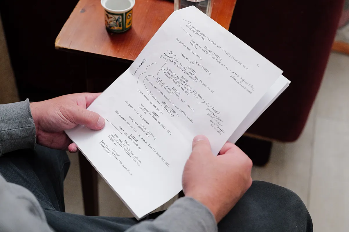 Person holding a typed script with handwritten notes, sitting at a wooden table with a cup.