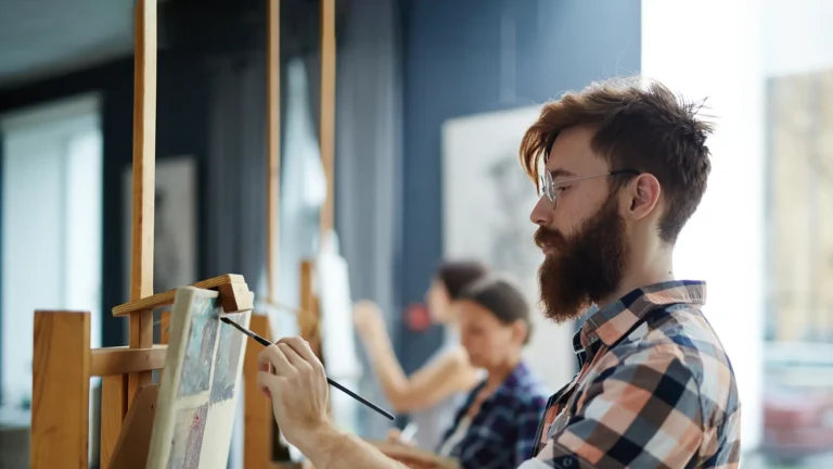 A bearded man wearing glasses and a checkered shirt paints on a canvas in an art class.