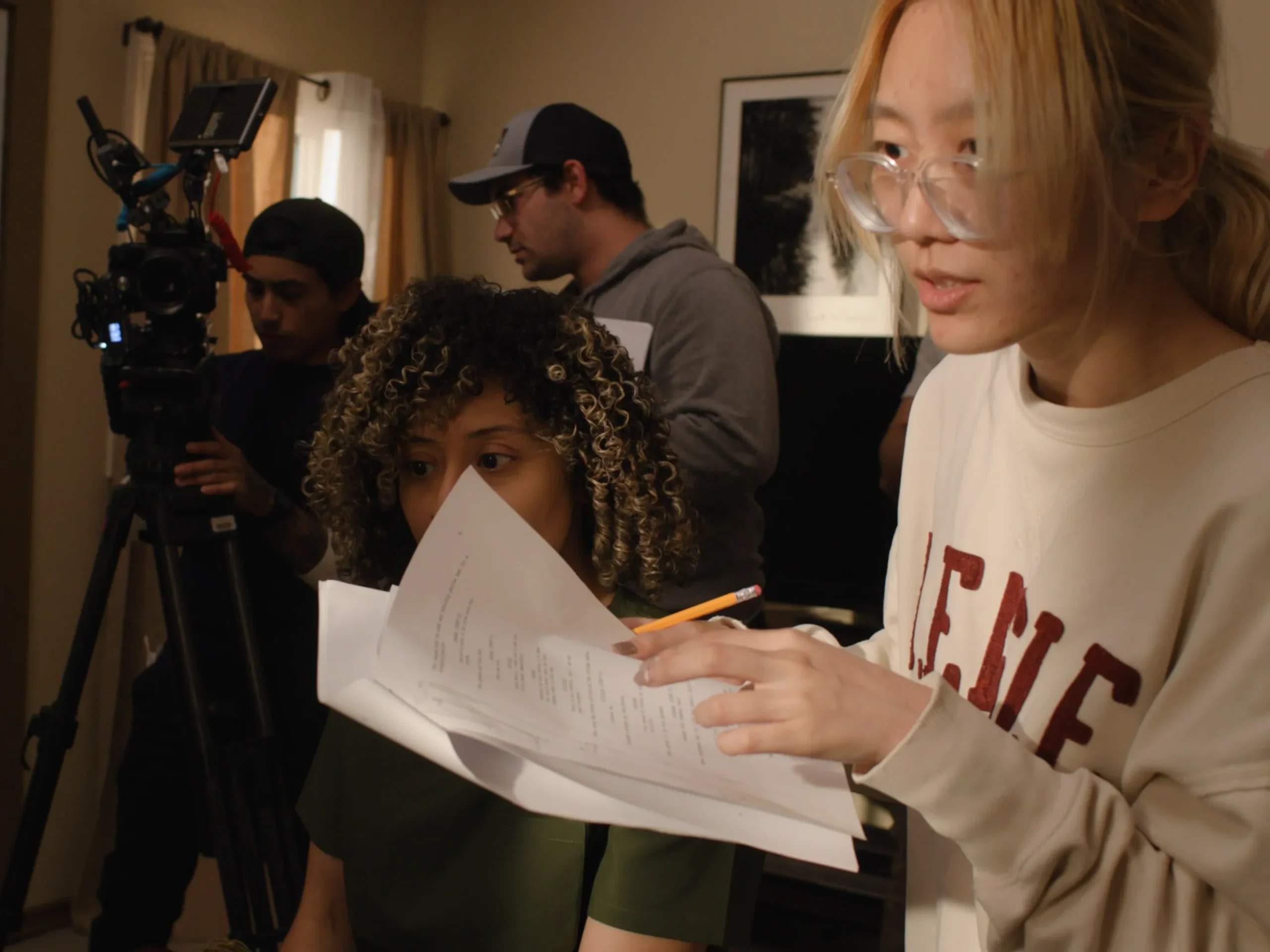 A woman with glasses holding a script, surrounded by film crew preparing for a shoot.