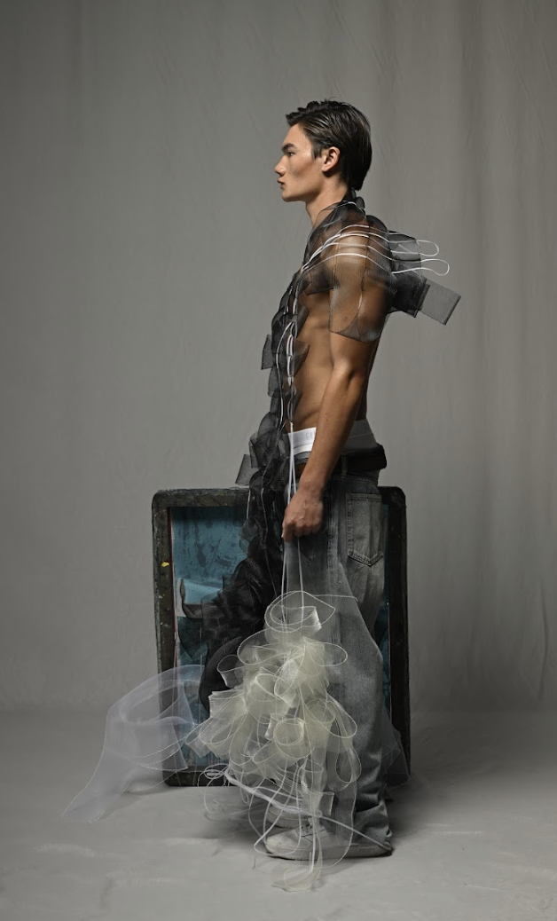 A shirtless young man with dark hair stands sideways next to a black bag and open blue suitcase.