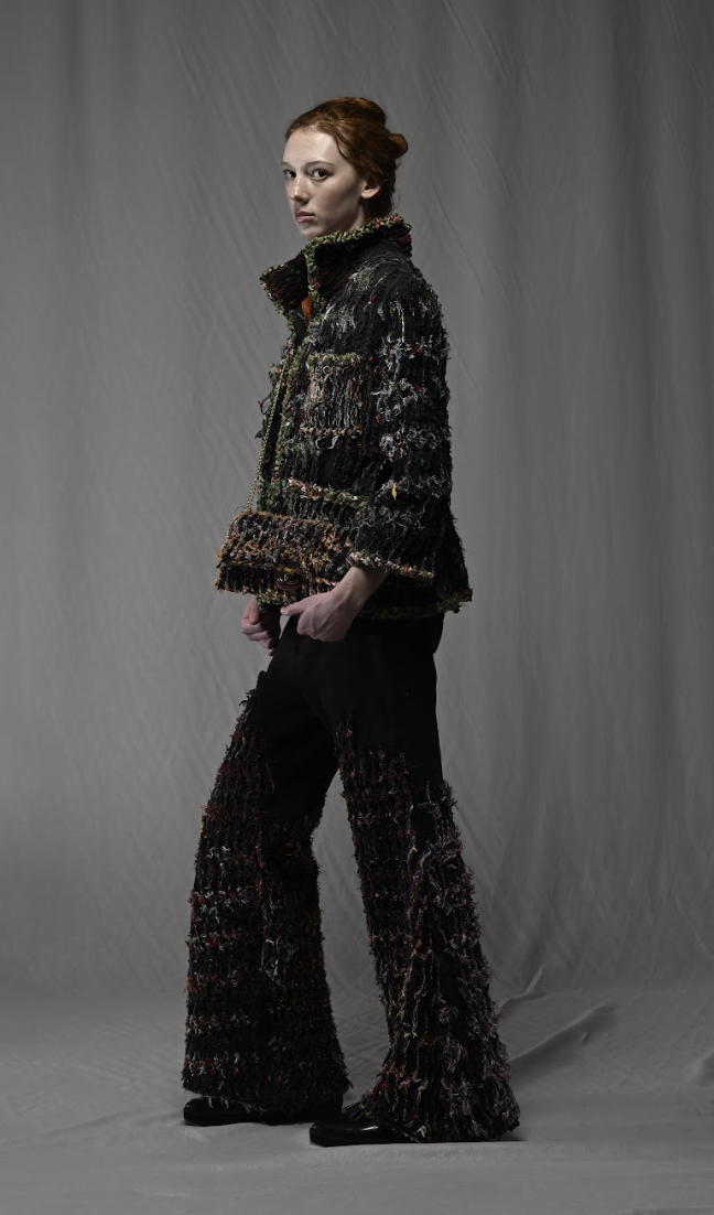 A woman with red hair styled in a bun, wearing a textured black jacket and matching wide-leg pants, standing against a gray backdrop.