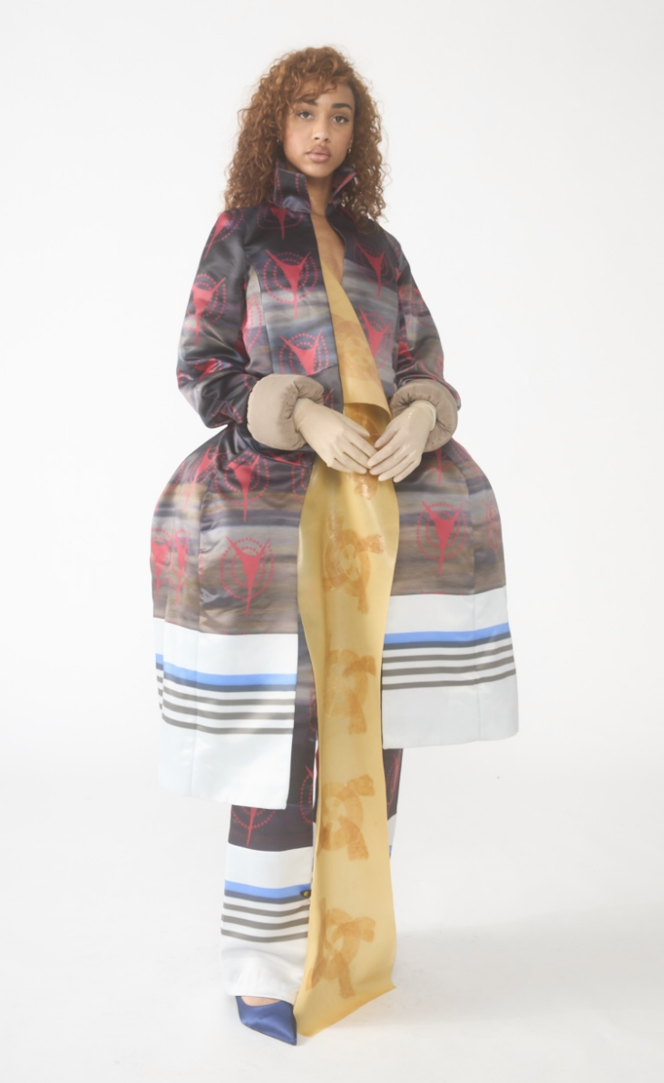 A woman standing with hands clasped, wearing a patterned coat and a long yellow scarf.