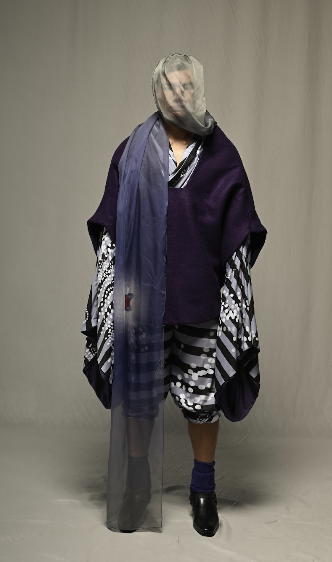 Person wearing layered eclectic clothing with a sheer face covering, standing against a plain background.