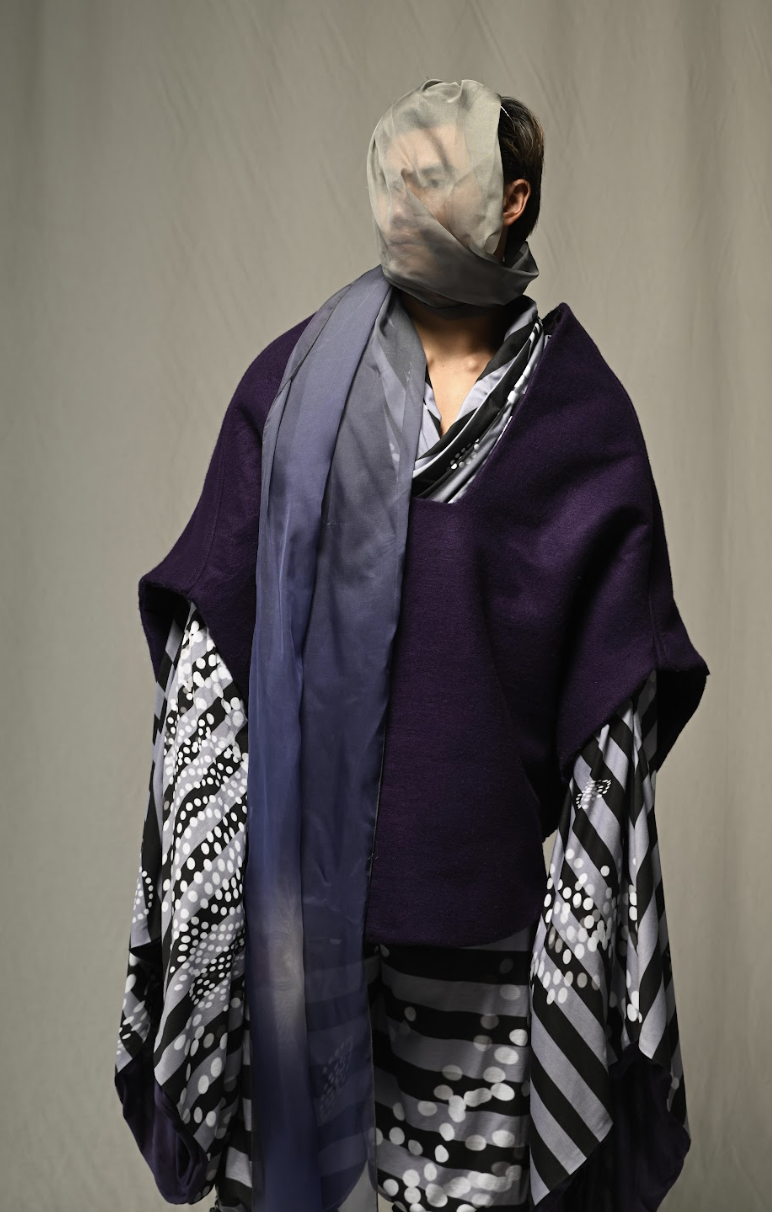 Person with face obscured by sheer fabric, wearing layered patterned and solid clothing.