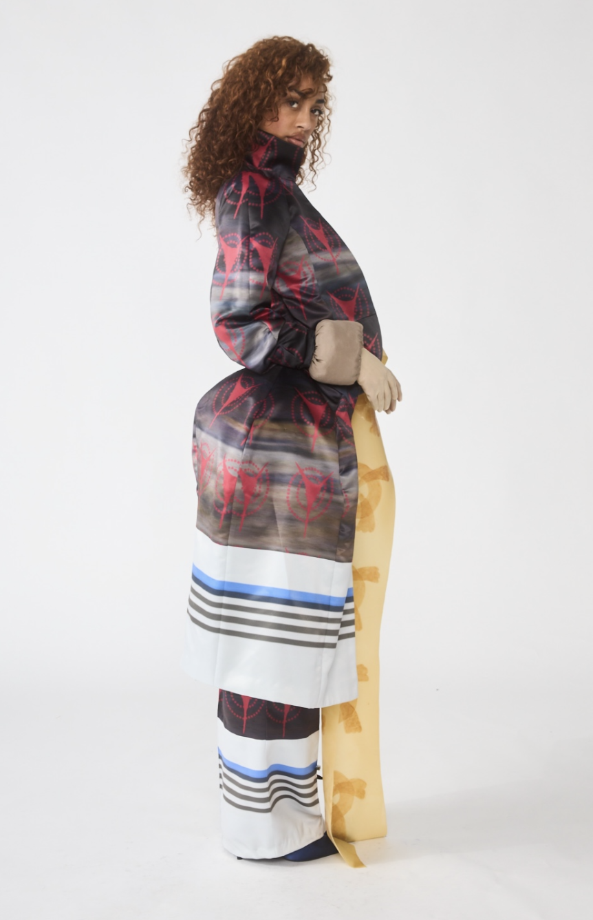 A woman stands sideways, wearing a colorful patterned coat, wide-leg pants, and holding a rolled-up yellow fabric.