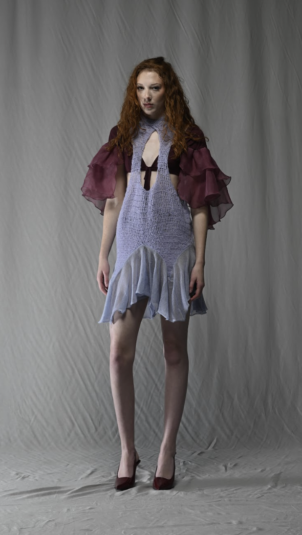 A woman with long, curly red hair wears a layered, ruffled dress in purple and blue tones.