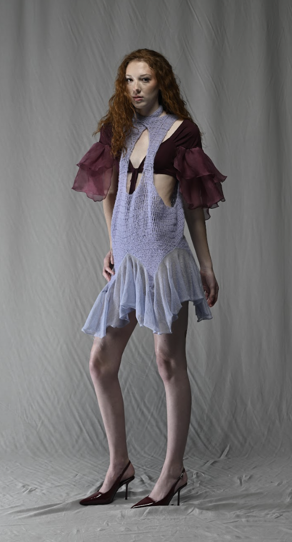 A woman with long, curly red hair wearing a layered dress and high heels, posing against a plain backdrop.