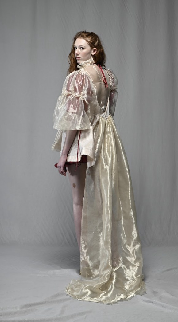 A woman in a layered, satin and sheer ruffled dress, standing against a plain backdrop.
