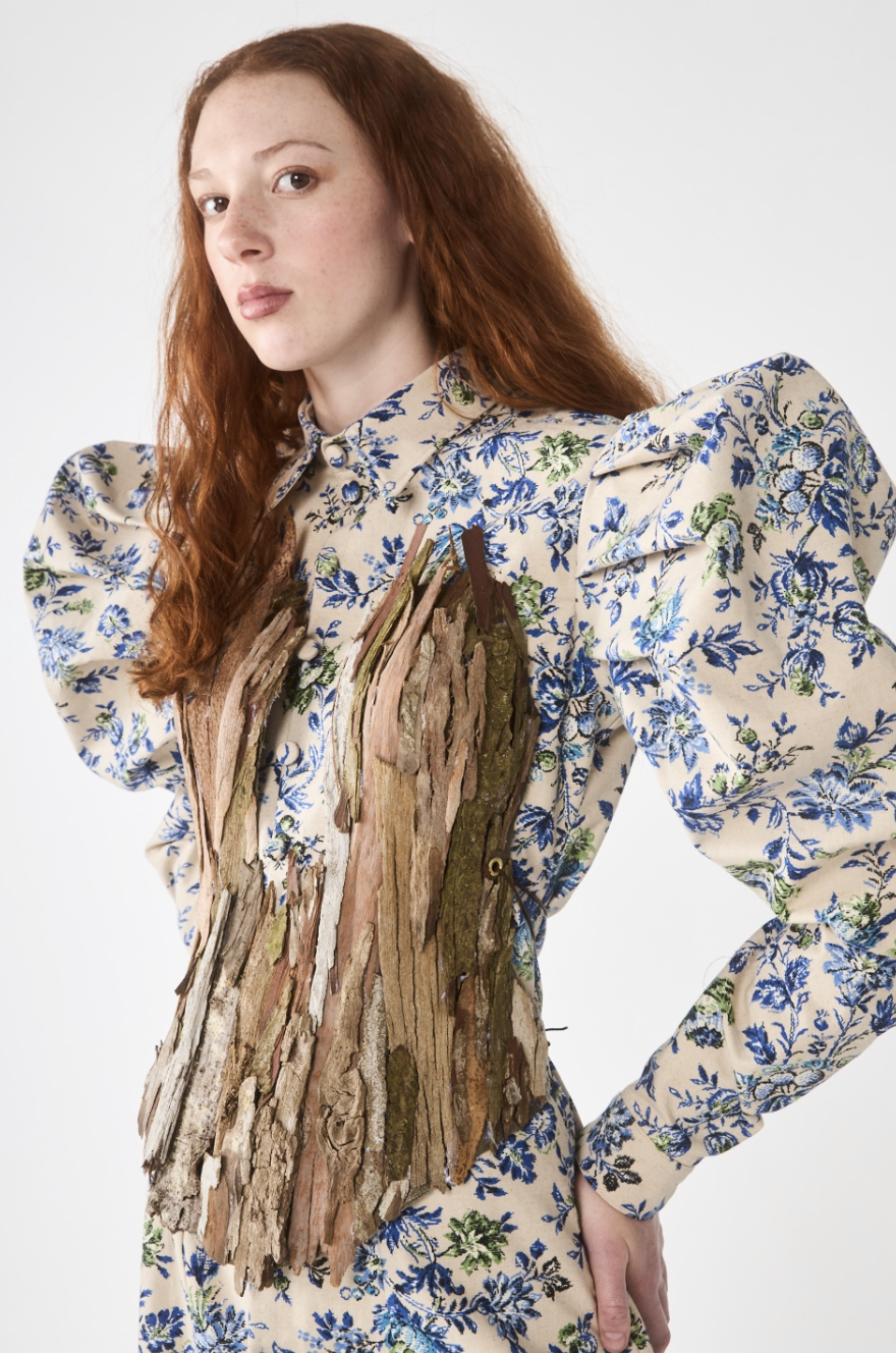 A woman with long red hair wearing a floral dress with puffed sleeves and a bark-covered piece of wood attached to her front.