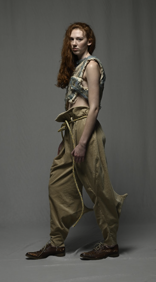 A woman with long red hair stands in a neutral pose, wearing oversized pants, a distressed top, and brown shoes.