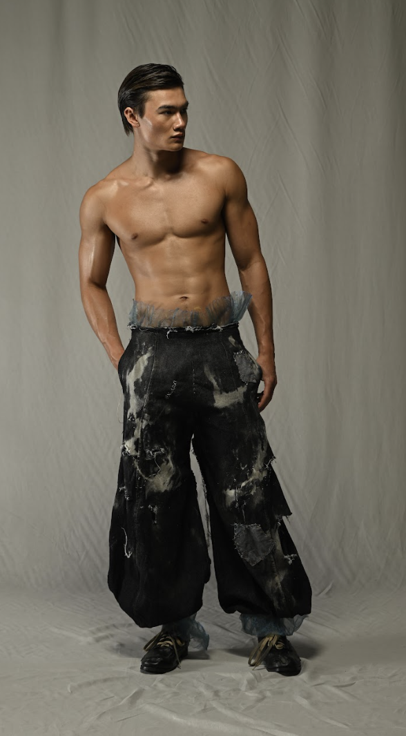 A shirtless man with black pants and sneakers, standing against a plain backdrop.