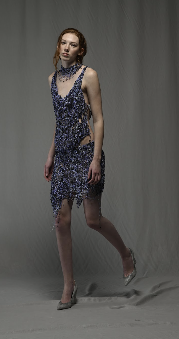 A woman in a damaged, shimmering blue dress with cutouts, standing on a gray backdrop.