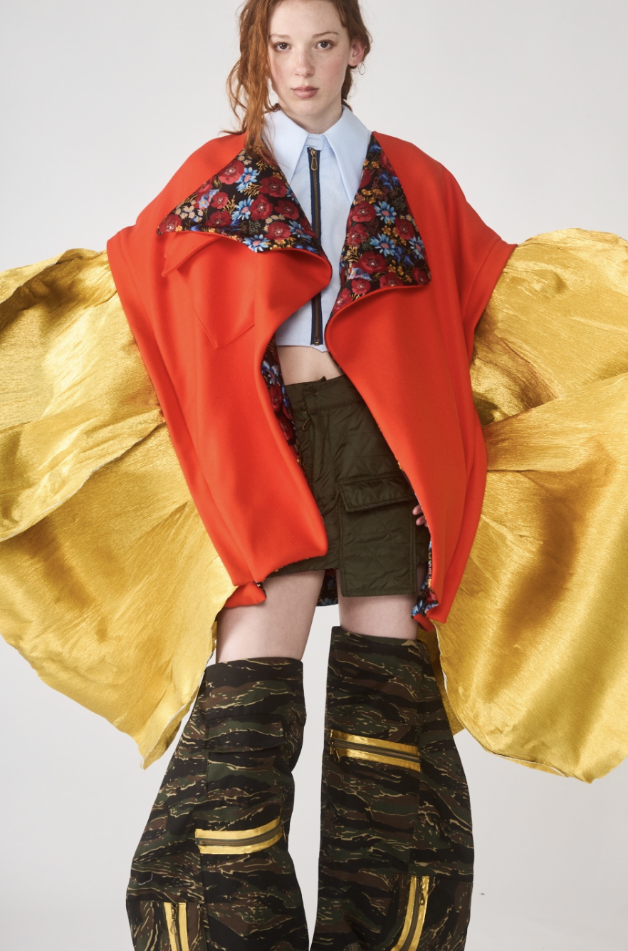 A woman wears a colorful layered outfit with a red coat, floral lining, black shorts, and camo-patterned boots.