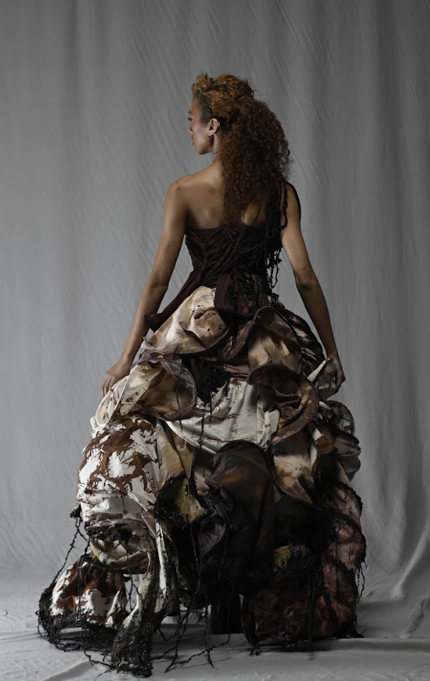 A woman in an elaborate, multi-textured gown with earthy colors, facing away from the camera