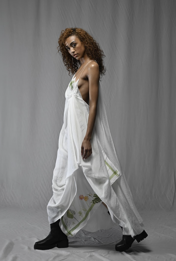 Woman wearing a flowing white dress with green and floral details, standing against a neutral background.