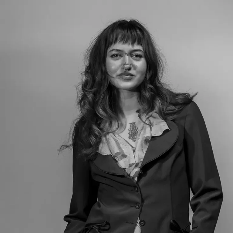 A woman with wavy hair and tattoos, wearing a blazer and floral blouse, looks at the camera.