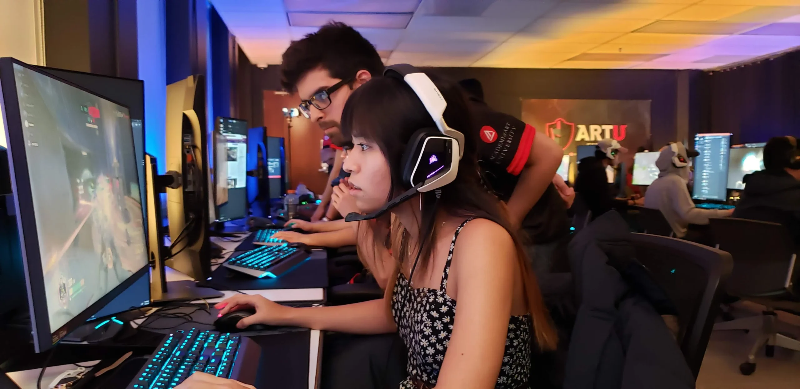 A woman in a headset playing a video game at a gaming event with others in the background.