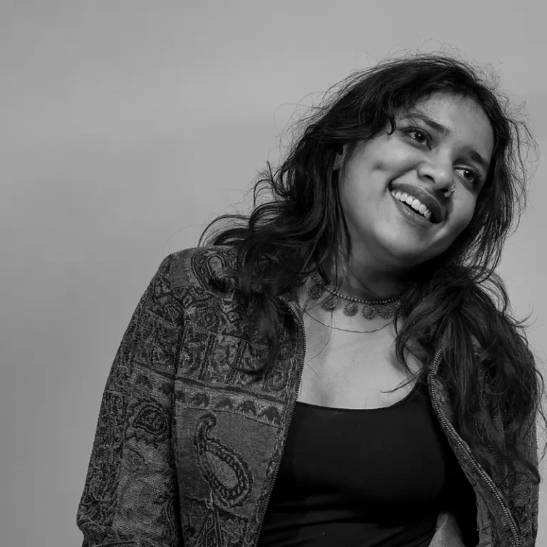 A smiling woman with wavy hair wearing a patterned jacket and choker necklace.