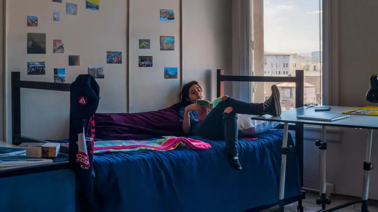 Girl lying on bed reading a book, with a window showing buildings outside.