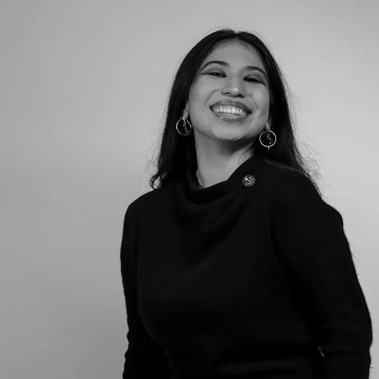 Smiling woman with long dark hair wearing earrings and a black top against a plain background.