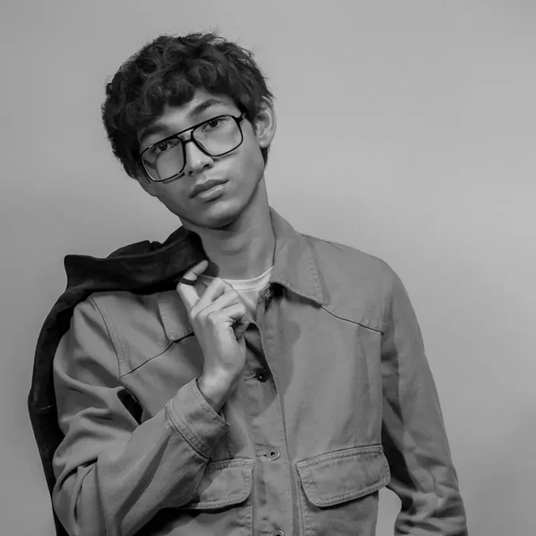 Young man with glasses holding a jacket over his shoulder in a neutral pose.