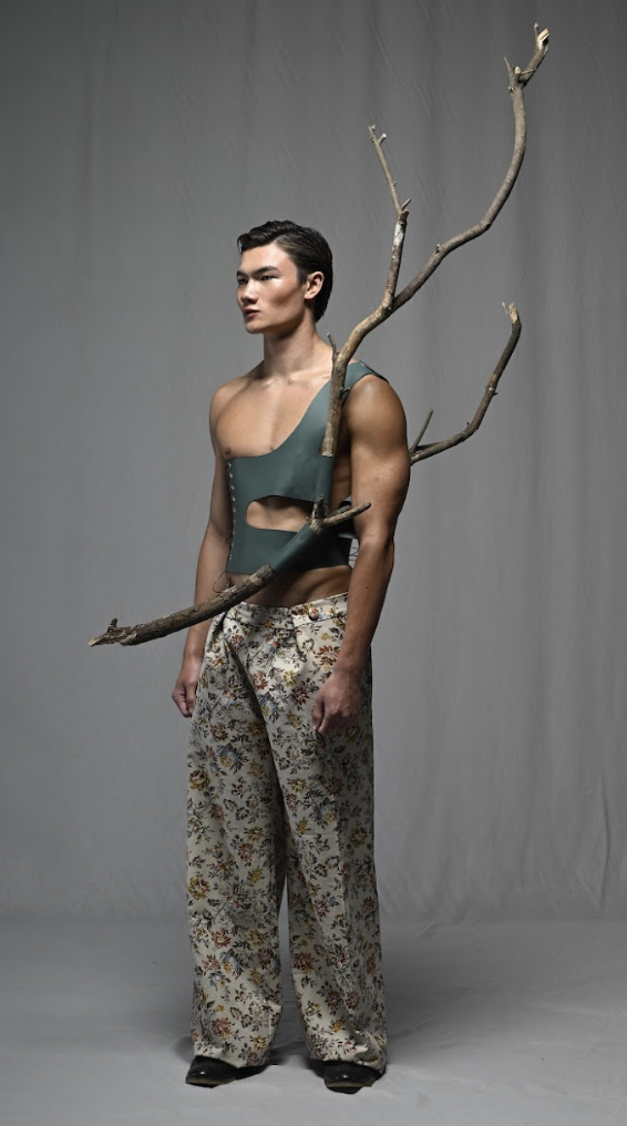 Man standing with a large tree branch across his shoulder, wearing floral pants and a cutout top.