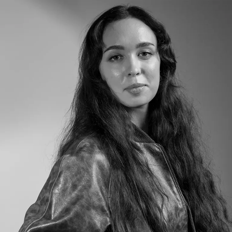 A woman with long wavy hair wearing a leather jacket looks at the camera.