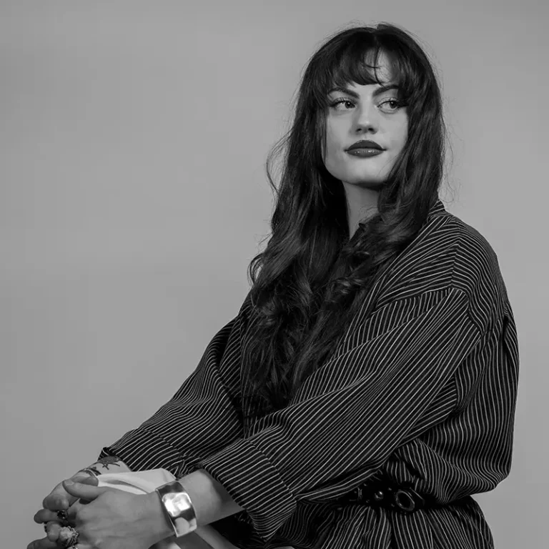A woman with long wavy hair and bold makeup, wearing a striped shirt and bracelet.